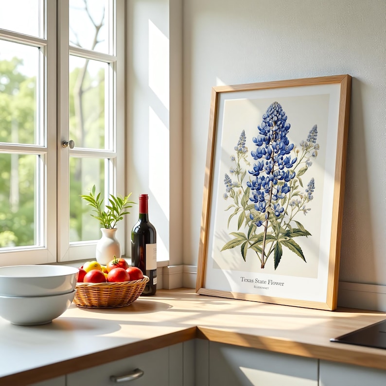 May include: A framed print of the Texas State Flower, a bluebonnet, in a wooden frame. The image is on a kitchen countertop next to a window, a bowl, a fruit basket, and a bottle of wine.
