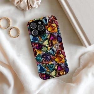 Puede incluir: Una funda de teléfono inteligente con un diseño vibrante de joyas. La funda presenta un mosaico de gemas coloridas y facetadas en tonos de azul, morado, amarillo y verde. La funda tiene un borde negro y un recorte para la cámara.