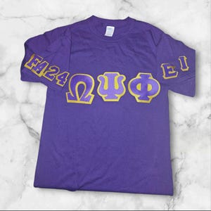 May include: Purple t-shirt with gold lettering on the sleeves and front. The sleeves say "F'24" and "E'1". The front says "ΩΨΦ".
