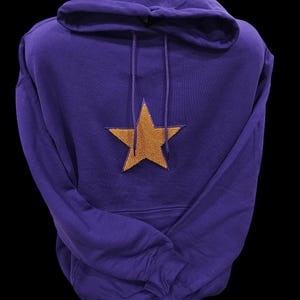 May include: A purple hooded sweatshirt with a gold star emblem on the front. The sweatshirt has a drawstring hood and a kangaroo pocket. The star is centered on the chest.