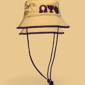 May include: Tan bucket hat with purple trim and embroidered Greek letters. The hat has a black adjustable chin strap. The hat has the Greek letters Omega, Psi, and Phi embroidered on the front.