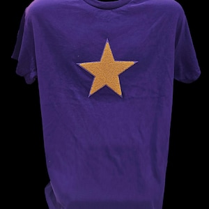 May include: A purple t-shirt with a large, gold star applique centered on the chest. The star has a textured appearance, contrasting with the smooth fabric of the shirt. The t-shirt has short sleeves and a crew neck.