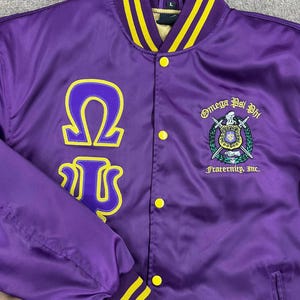 May include: A purple satin jacket with yellow accents. The jacket features the Greek letters Omega and Psi, along with the words "Omega Psi Phi Fraternity, Inc." on the front. The collar and cuffs have yellow stripes.