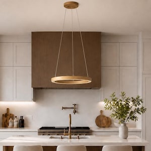 May include: A modern kitchen with white cabinetry, a wooden range hood, and a central island with three beige bar stools. A gold-toned circular pendant light hangs above the island. A vase of greenery sits on the island.
