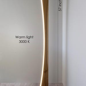 May include: A wooden floor lamp with a curved design, featuring a warm light (3000 K) that runs along its length. The lamp is 144 cm tall, with a base of 35 cm by 24.5 cm.