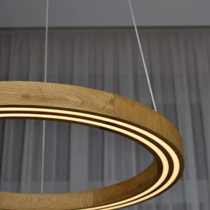 May include: A modern, circular pendant light fixture. The light has a wooden frame with a light strip on the inside. The light is suspended from the ceiling by thin white cords. The wood has a natural, light brown color.