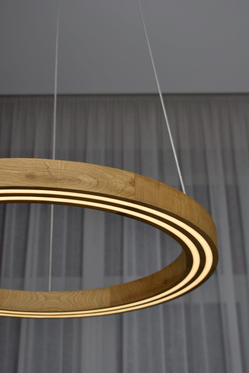 May include: A modern, circular pendant light fixture. The light has a wooden frame with a light strip on the inside. The light is suspended from the ceiling by thin white cords. The wood has a natural, light brown color.