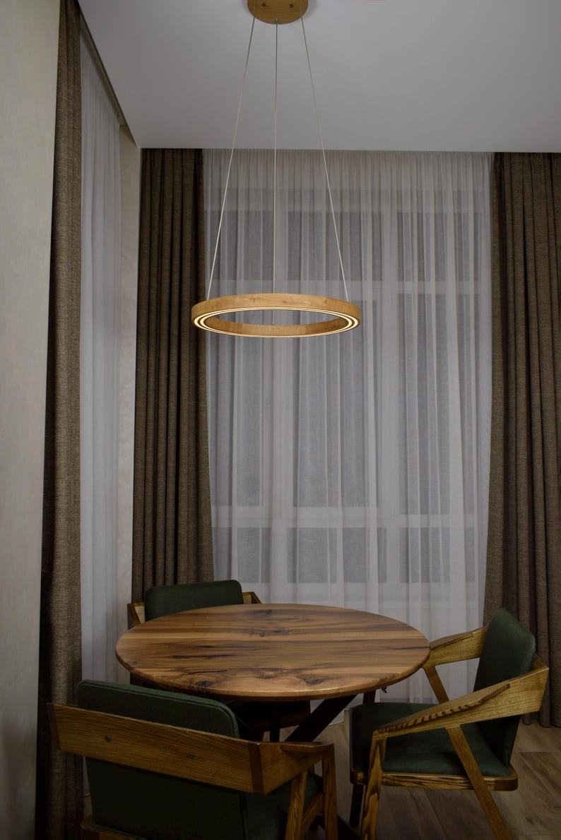 May include: A modern wooden ring pendant light fixture hangs above a round wooden table and chairs. The light has a warm, inviting glow. The room has brown curtains and sheer white curtains.