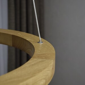 May include: Close-up of a light-colored wooden ring suspended by a thin, silver-colored cable. The ring has a smooth, curved surface and appears to be made of solid wood. The background is a blurred gray.