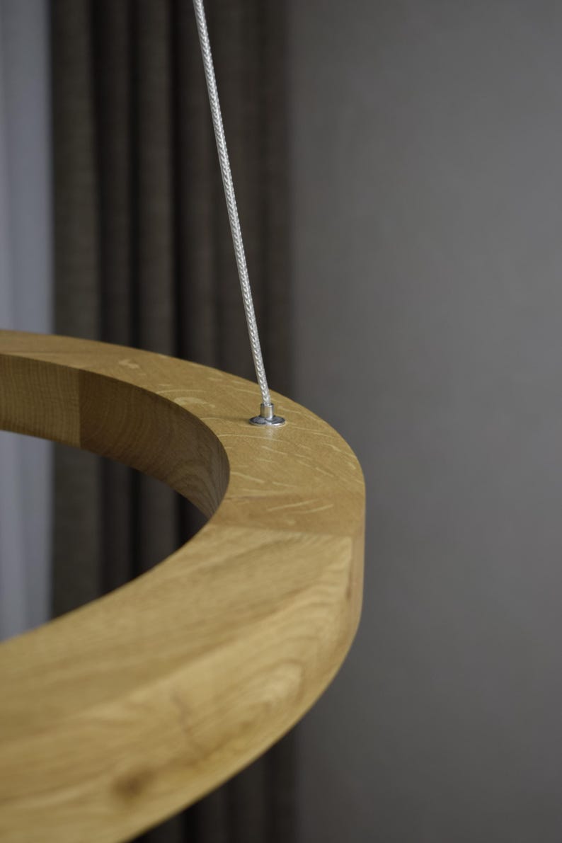 May include: Close-up of a light-colored wooden ring suspended by a thin, silver-colored cable. The ring has a smooth, curved surface and appears to be made of solid wood. The background is a blurred gray.