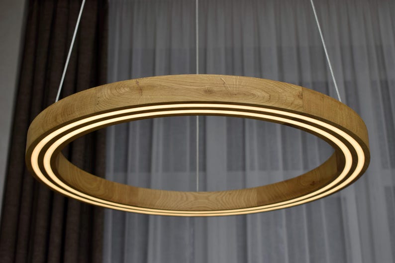 May include: A modern, circular pendant light fixture. The light has a wooden frame with two parallel, illuminated bands of light. The fixture hangs from two thin white cords against a neutral background.