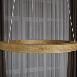 May include: A circular wooden pendant light fixture. The light is suspended from the ceiling by three thin white cords. The light has a natural wood grain finish and is positioned in front of a window with sheer white curtains and brown drapes.