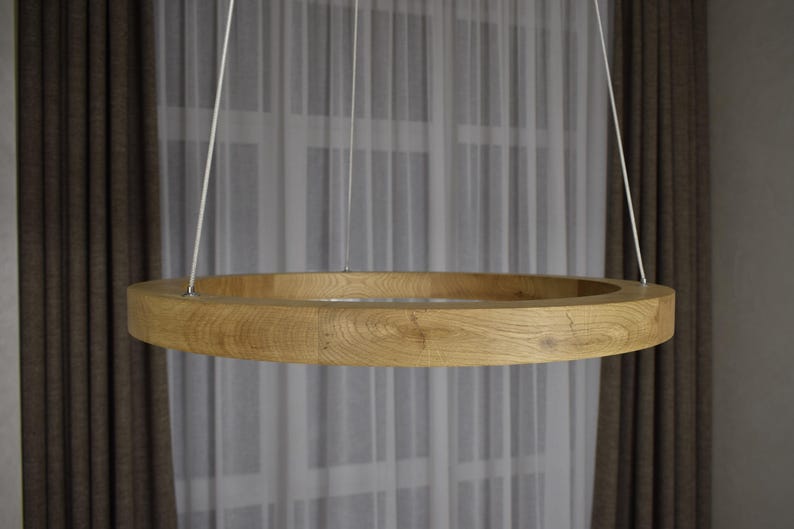 May include: A circular wooden pendant light fixture. The light is suspended from the ceiling by three thin white cords. The light has a natural wood grain finish and is positioned in front of a window with sheer white curtains and brown drapes.