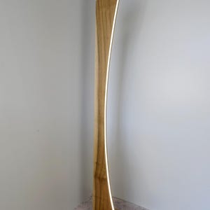 May include: A contemporary wooden floor lamp with a curved design. The lamp has a warm, natural wood finish and a vertical, illuminated strip of light. The base is rectangular, and the lamp stands tall against a neutral wall.