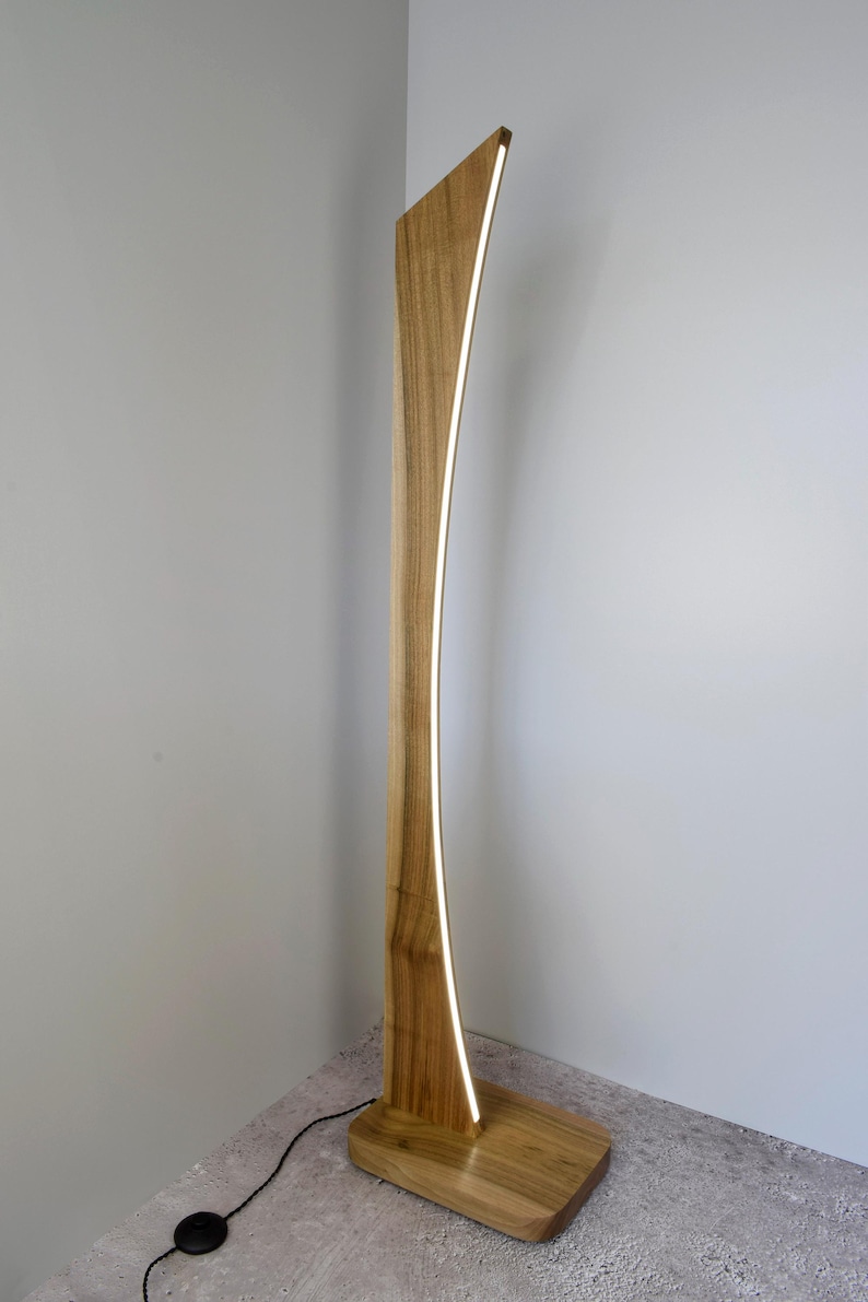 May include: A contemporary wooden floor lamp with a curved design. The lamp has a warm, natural wood finish and a vertical, illuminated strip of light. The base is rectangular, and the lamp stands tall against a neutral wall.