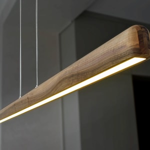 Può includere: Una lampada a sospensione moderna con un corpo in legno lungo e rettangolare e una striscia luminosa LED integrata. Il legno presenta una venatura naturale e un colore marrone caldo.