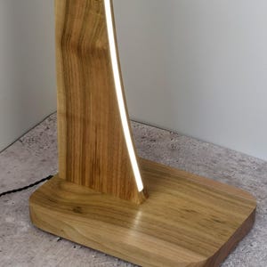 May include: A wooden floor lamp with a rectangular base and a tall, curved wooden support. A bright, white light strip runs along the inside curve of the support. The lamp is made of light-coloured wood and sits on a concrete floor.