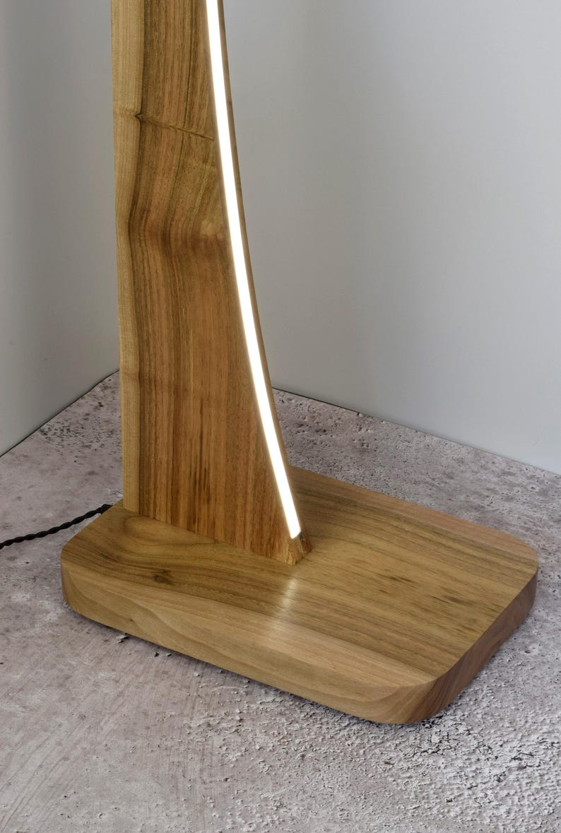 May include: A wooden floor lamp with a rectangular base and a tall, curved wooden support. A bright, white light strip runs along the inside curve of the support. The lamp is made of light-coloured wood and sits on a concrete floor.
