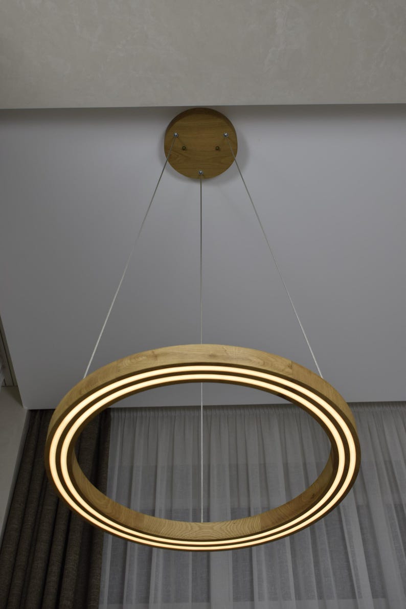 May include: A modern, circular pendant light fixture. The light features a wooden ring with two parallel, illuminated bands. The fixture is suspended from the ceiling by thin cables, with a wooden ceiling mount. The light is a warm, inviting color.