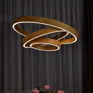 May include: A circular pendant light fixture constructed from light-toned wood with a visible grain pattern. The fixture features three interlocking rings of varying sizes, each equipped with an integrated LED strip that emits a warm white glow.