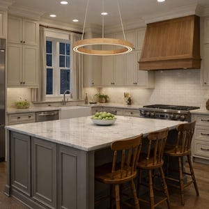 May include: A well-lit kitchen with a large island and wooden bar stools. A wooden circular light fixture hangs above the island. The kitchen features light-colored cabinets, a marble countertop, and a wooden range hood.