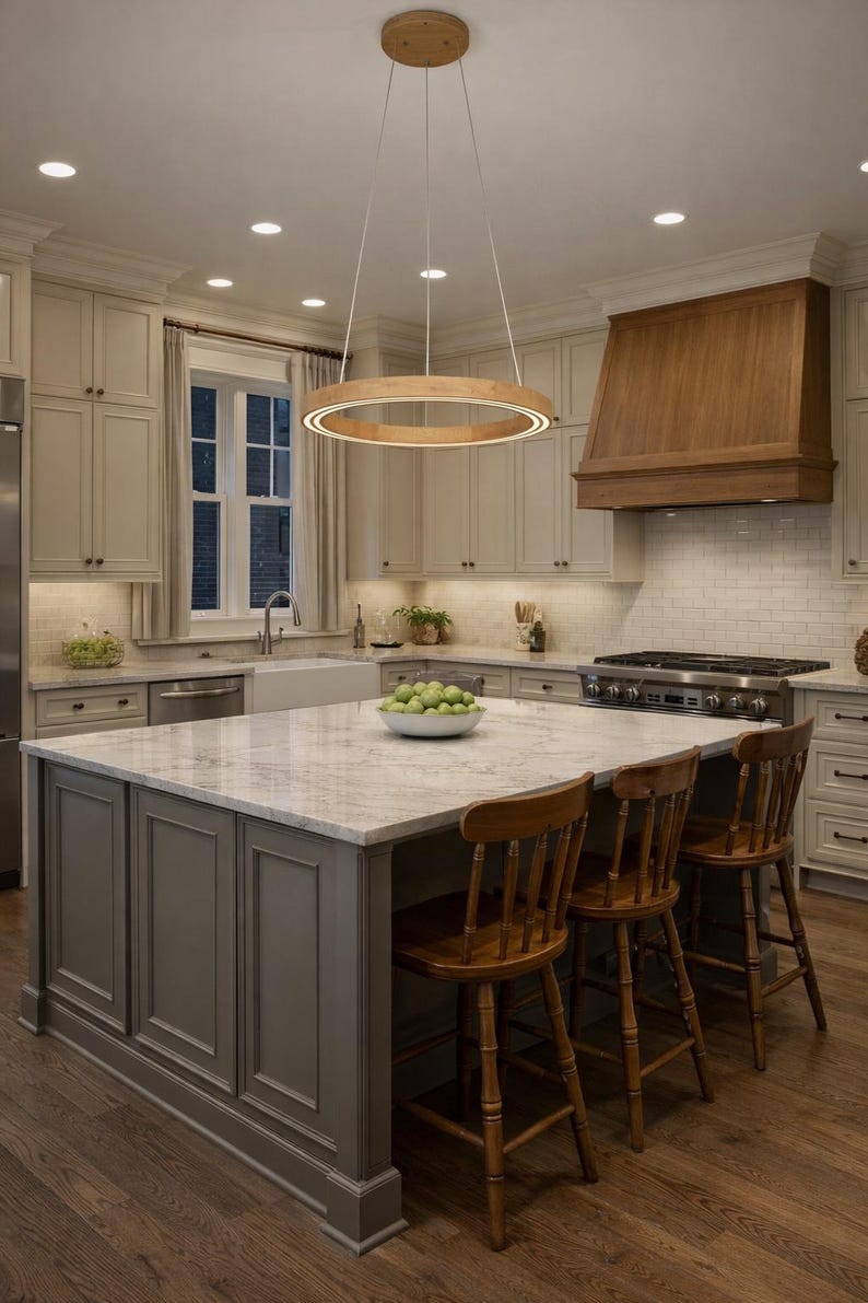 May include: A well-lit kitchen with a large island and wooden bar stools. A wooden circular light fixture hangs above the island. The kitchen features light-colored cabinets, a marble countertop, and a wooden range hood.