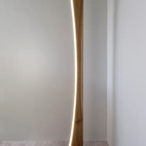 May include: A contemporary wooden floor lamp with a curved design. The lamp has a natural wood finish and a vertical LED light strip. The base is rectangular, and the lamp is plugged into an electrical outlet. The height is approximately 170 cm.