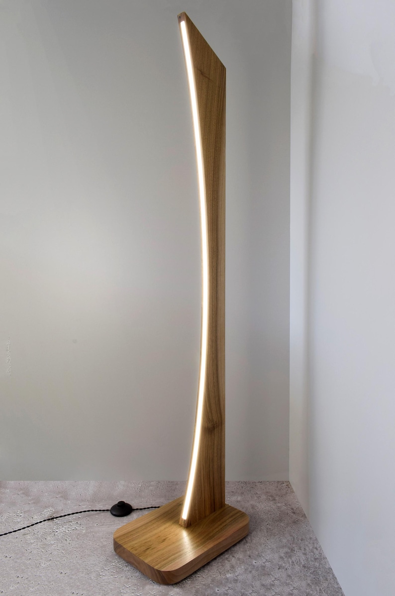 May include: A contemporary wooden floor lamp with a curved design. The lamp has a natural wood finish and a vertical LED light strip. The base is rectangular, and the lamp is plugged into an electrical outlet. The height is approximately 170 cm.