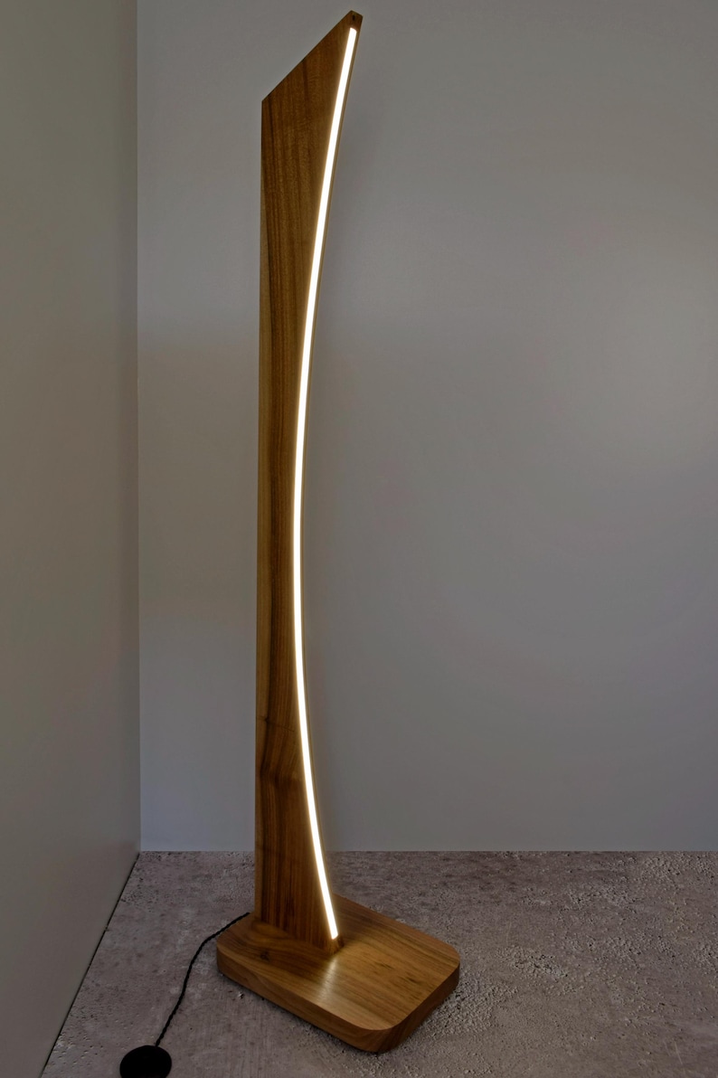 May include: A contemporary wooden floor lamp with a curved design. The lamp has a warm, natural wood finish and a bright, vertical LED light strip. The lamp is a stylish addition to any living space. The lamp's height is approximately 170 cm.