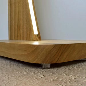 May include: A contemporary wooden lamp featuring a light wood base and a vertical illuminated strip. The base is a curved, rectangular form, supported by clear plastic feet. The design of the lamp is minimalist.