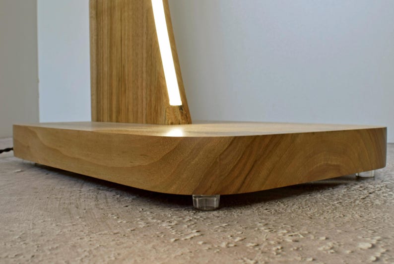 May include: A contemporary wooden lamp featuring a light wood base and a vertical illuminated strip. The base is a curved, rectangular form, supported by clear plastic feet. The design of the lamp is minimalist.