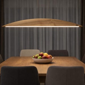 Peut inclure: Une longue suspension en bois clair et incurvé, fixée par de fins câbles au-dessus d'une table à manger rectangulaire en bois. Le luminaire présente une surface supérieure arquée et lisse avec une source lumineuse linéaire blanche.