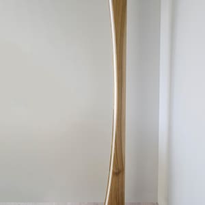 May include: A contemporary wooden floor lamp with a curved design. The lamp has a light strip running along its length and a rectangular base. The wood has a natural finish, and the lamp is plugged into a wall socket.