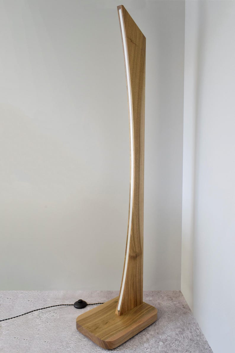 May include: A contemporary wooden floor lamp with a curved design. The lamp has a light strip running along its length and a rectangular base. The wood has a natural finish, and the lamp is plugged into a wall socket.