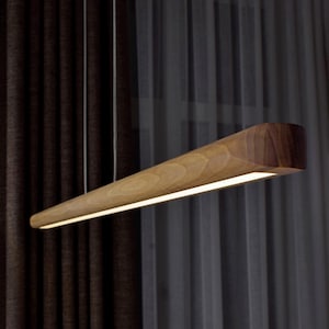 Peut inclure: Un luminaire suspendu moderne en bois. La lampe a une forme rectangulaire effilée avec une bande lumineuse en bas. Le bois a un grain naturel et une couleur marron chaud. La lampe est suspendue à de fins fils argentés.