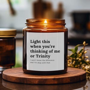 May include: A brown glass candle jar with a white label that reads "Light this when you're thinking of me or Trinity. I won't know the difference and I'm okay with that."