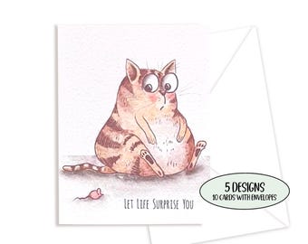 May include: A greeting card featuring a watercolor illustration of a round, brown tabby cat with wide eyes and a small pink mouse. The text "LET LIFE SURPRISE YOU" is printed below the cat. An oval badge states "5 DESIGNS 10 CARDS WITH ENVELOPES".