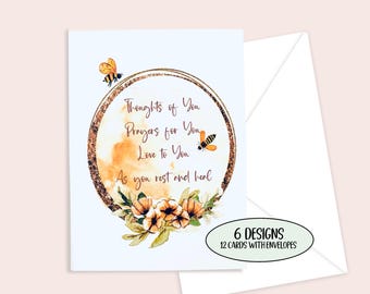 May include: A set of greeting cards with a white background. The card features a gold-colored circle with the text "Thoughts of You, Prayers for You, Love to You, As you rest and heal." Two bees and floral accents are also included. The set includes 6 designs and 12 cards with envelopes.