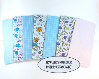 May include: A set of seven traveler's notebook inserts with various covers including blue grid patterns, a floral print, a cartoon cat and mouse design, a botanical pattern with purple and yellow fruits, and a light pink grid pattern.