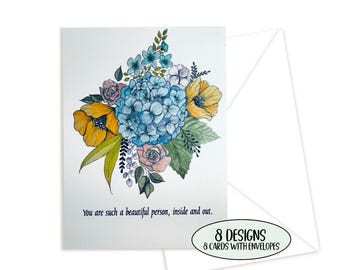 May include: A white greeting card featuring a watercolor illustration of a blue hydrangea, yellow poppies, and green leaves. The text reads, You are such a beautiful person, inside and out. The set includes 8 designs, 8 cards, and 8 envelopes.