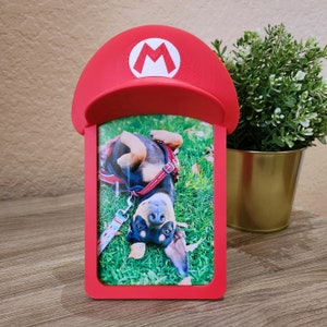 May include: Red picture frame shaped like a Super Mario mushroom with a photo of a black and brown dachshund wearing a red harness and lying on its back in green grass.