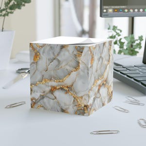 May include: A white and gold marble patterned notepad with a white top sheet. The notepad is sitting on a white surface.
