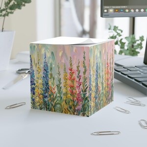 May include: A cube-shaped notepad with a floral painting. The artwork features colorful wildflowers in shades of blue, yellow, pink, and green against a soft pastel background. The notepad sits on a white surface with office supplies.