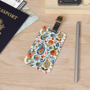 May include: A white luggage tag with a colorful paisley pattern in shades of blue, orange, yellow, and green. The tag has a black strap and a gold buckle.