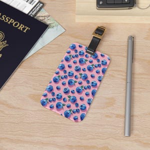 May include: Pink luggage tag with a repeating pattern of blue blueberries on a pink background. The tag has a brown leather strap with a metal buckle.