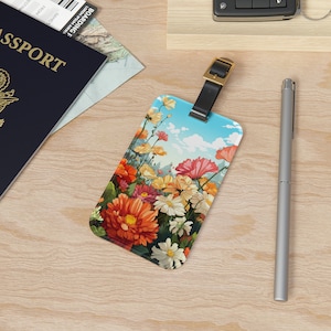 May include: A luggage tag with a colorful floral design. The tag features a variety of flowers in shades of pink, orange, yellow, and white, set against a blue sky with white clouds. The tag has a black strap with a gold buckle.