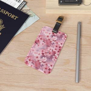 May include: A rectangular luggage tag with a floral cherry blossom design in shades of pink and white. The tag has a black leather strap with a gold buckle. A silver pen and a passport are also visible.
