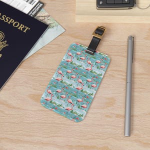 May include: A blue luggage tag with a pattern of pink flamingos on a blue background. The tag has a brown leather strap and a gold metal buckle.
