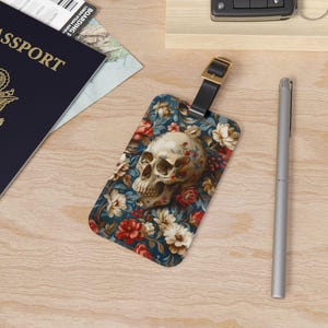 May include: A luggage tag with a skull and floral design. The skull is white with red and blue accents. The flowers are red, white, and blue. The tag is made of a durable material and has a leather strap.
