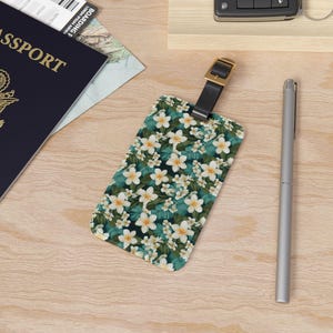 May include: A luggage tag with a tropical floral pattern. The tag is green with white flowers and has a black leather strap with a gold buckle.